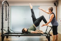 Concentrated male in sportswear doing exercises on pilates reformer during training under supervision of female instructor in protective respirator during coronavirus pandemic — Stock Photo