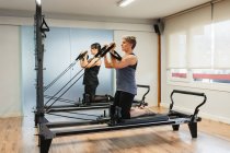 Seitenansicht fokussierter Sportlerinnen in Aktivkleidung, die Übungen auf Pilates-Geräten machen und Muskeln mit Metallwiderstandsgeräten pumpen — Stockfoto