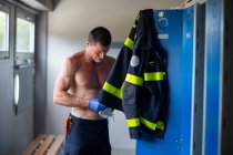 Ernsthafter Feuerwehrmann mit nacktem Oberkörper und in Latexhandschuhen steht mit Atemschutzmaske in der Nähe des Spind am Feuerwehrhaus während der Arbeitsvorbereitung — Stockfoto