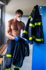 Ernsthafter Feuerwehrmann mit nacktem Oberkörper und in Latexhandschuhen steht mit Atemschutzmaske in der Nähe des Spind am Feuerwehrhaus während der Arbeitsvorbereitung — Stockfoto