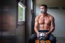 Strong mature male firefighter with naked torso sitting on bench in mask and holding a helmet while looking away — Stock Photo
