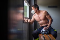 Strong mature male firefighter with naked torso sitting on bench in mask and holding a helmet while looking away on small windows — Stock Photo
