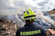 Обратный вид отважного пожарного в защитной форме, стоящего со шлангом и тушащего пожар на свалке в горах — стоковое фото