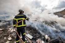 Back view of brave fireman in protective uniform standing with hose and extinguishing fire on dump in mountains — Stock Photo