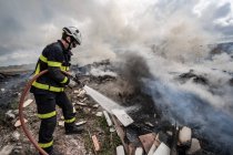 Vista laterale di coraggioso pompiere in uniforme protettiva in piedi con tubo flessibile ed estinguente incendio in discarica in montagna — Foto stock