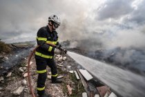 Vista lateral do bravo bombeiro em uniforme de proteção em pé com mangueira e extinguindo o fogo no despejo nas montanhas — Fotografia de Stock