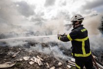 Seitenansicht des tapferen Feuerwehrmannes in Schutzuniform, der mit Schlauch steht und Feuer auf Müllkippe in den Bergen löscht — Stockfoto
