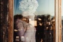 Улыбающаяся женщина в уютной одежде, стоящая у окна и закрывающая лицо цветком гортензии, глядя в камеру — стоковое фото