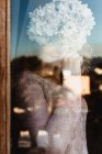 Mulher sorridente em roupas aconchegantes de pé perto da janela e cobrindo o rosto com flor de hortênsia enquanto olha para a câmera — Fotografia de Stock