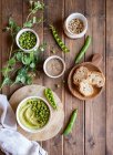 Zusammensetzung von oben mit Schüssel mit Humus aus grünen Erbsen auf Holztisch mit Zutaten für Rezept und Brotscheiben angeordnet — Stockfoto