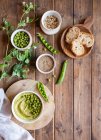 Composição vista superior com tigela com hummus feito com ervilha verde dispostos em mesa de madeira com ingredientes para receitas e fatias de pão — Fotografia de Stock