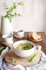 Состав с миской с хумусом, сделанный из зеленого горошка на деревянном столе с ингредиентами для рецепта и ломтиками хлеба — стоковое фото