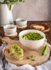 Komposition mit Schüssel mit Humus aus grünen Erbsen auf Holztisch mit Zutaten für Rezept und Brotscheiben — Stockfoto