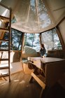 Femme en tenue décontractée assise à table dans une maison futuriste en bois située au camping en forêt par une journée ensoleillée — Photo de stock