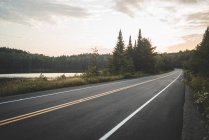 Route asphaltée à proximité d'un lac paisible et d'une forêt verdoyante contre un ciel nuageux au coucher du soleil dans le parc national de la Mauricie au Québec, Canada — Photo de stock