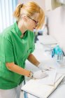 Focused adult woman in glasses and uniform working in dental hospital and writing on paper card at table — Stock Photo