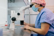Visão lateral da mulher de uniforme médico e máscara usando microscópio moderno para examinar células humanas enquanto trabalhava no laboratório da clínica moderna — Fotografia de Stock