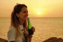 Улыбающаяся юная леди с бутылкой пива во время заката на берегу моря — стоковое фото