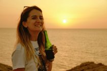 Happy young female traveler in casual clothes and sunglasses holding glass beer bottle while relaxing during sunset on coast and looking at camera in Ibiza — Stock Photo