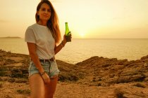 Усміхнена молода леді з пляшкою пива під час заходу сонця на березі моря — стокове фото