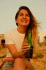 Sorridente giovane signora con bottiglia di birra durante il tramonto in riva al mare — Foto stock