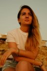 Basso angolo di donna in abiti casual seduto sulla spiaggia rocciosa e guardando lontano sognante durante il tramonto a Ibiza — Foto stock