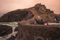 Visão traseira de turista feminina irreconhecível em roupas quentes caminhando em direção à ponte da capela enquanto viaja na ilha de San Juan de Gaztelugatxe, na Espanha — Fotografia de Stock