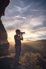 Vista laterale del giovane viaggiatore maschio in abiti casual in piedi sulla collina e scattare foto di paesaggi spettacolari della catena montuosa al tramonto — Foto stock