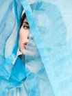 Очаровательная модель в прозрачной синей блузке и покрывающей лицо текстилем, глядя на камеру в студии — стоковое фото