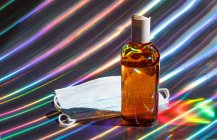 Mascarilla médica protectora y botella de gel desinfectante marrón sobre fondo holográfico brillante - foto de stock