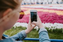 Gesichtslose Reisende in Jeanshemd und Sonnenbrille fotografieren bunte große Blumenbeete auf dem Handy, während sie in der Nähe des Zauns stehen und auf den Bildschirm schauen — Stockfoto