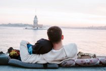Visão traseira do casal amoroso anônimo abraçando sentado em almofadas à beira-mar e desfrutando do pôr do sol maravilhoso durante a data romântica em Istambul — Fotografia de Stock
