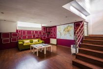 Современная просторная гостиная с деревянным полом и лестницей, обставленная ярко-зеленым диваном и украшенная геометрическими элементами на красных стенах — стоковое фото