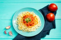 Vue de dessus de assiette en céramique bleue avec pâtes et sauce tomate décorée de persil et basilic servi entre gousses d'ail et couple de tomates sur fond bleu clair — Photo de stock