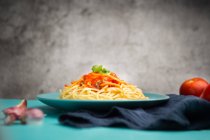 Голубая керамическая тарелка с макаронами и томатным соусом, украшенная петрушкой и базиликом, подается между зубчиком чеснока и парой помидоров на светло-голубом фоне — стоковое фото