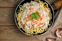 From above tasty spaghetti with ham slices and mushrooms in creamy sauce cooked in pan and placed on wooden cutting board at a wooden table with garlic and linen fabric aside — Stock Photo