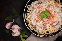Composition of tasty spaghetti with ham slices and mushrooms in creamy sauce cooked in pan and placed on wooden cutting board at dark table with garlic and gray linen fabric aside — Stock Photo