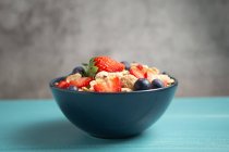 Leckere Frühstücksschüssel mit Cornflakes mit Erdbeeren und Blaubeeren auf Schneidebrett gelegt und mit Leinentuch und Beeren um Teller auf blauem Holztisch mit grauem Hintergrund dekoriert — Stockfoto