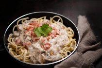 Composition of tasty spaghetti with ham slices and mushrooms in creamy sauce cooked in pan and placed on wooden cutting board at dark table with garlic and gray linen fabric aside — Stock Photo