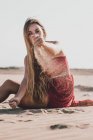 Jovem atraente com longos cabelos loiros vestindo elegante vestido vermelho sentado na costa e derramando areia da mão enquanto olha para a câmera — Fotografia de Stock