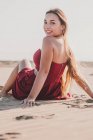 Привлекательная молодая леди с длинными светлыми волосами в стильном красном платье сидит на берегу, глядя в камеру — стоковое фото