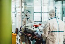 Невпізнавані професійні лікарі в захисній формі та масках піклуються про пацієнта з вірусною інфекцією, стоячи в операційній кімнаті в сучасній лікарні — стокове фото