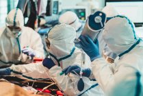 Gruppe professioneller Ärzte mit Schutzmasken und Anzügen, die in der Nähe des Operationstisches mit Geräten stehen und sich auf die Operation in einer modernen Klinik vorbereiten — Stockfoto