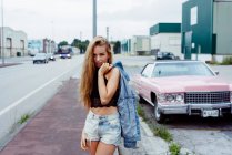 Чуттєва блондинка стоїть на тротуарі і дивиться на камеру біля класичного рожевого автомобіля. — стокове фото