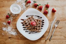 Soft sweet waffles and strawberries served on plate with yummy chocolate syrup and coconut flakes on wooden table near silverware — Stock Photo