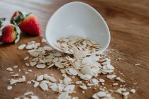 Bowl of healthy almond chips spilled on wooden table near ripe strawberries in kitchen — Stock Photo