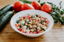 Von oben Schüssel mit gesunden Gurken und Tomatensalat mit Kichererbsen und Petersilie auf Holztisch neben Gabel und Messer gelegt — Stockfoto