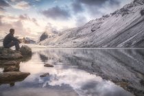 Vista lateral del macho adulto sentado en piedra cerca del lago tranquilo y la montaña nevada y admirando el cielo nublado al atardecer mientras visita el Parque Nacional Suizo en Suiza - foto de stock