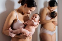 Mulher encantada em roupa interior abraçando bebê nu perto do espelho enquanto se inclina na parede e sorrindo — Fotografia de Stock