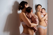 Entzückte Frau in Unterwäsche kuschelt nacktes Baby neben Spiegel, während sie sich an Wand lehnt und lächelt — Stockfoto